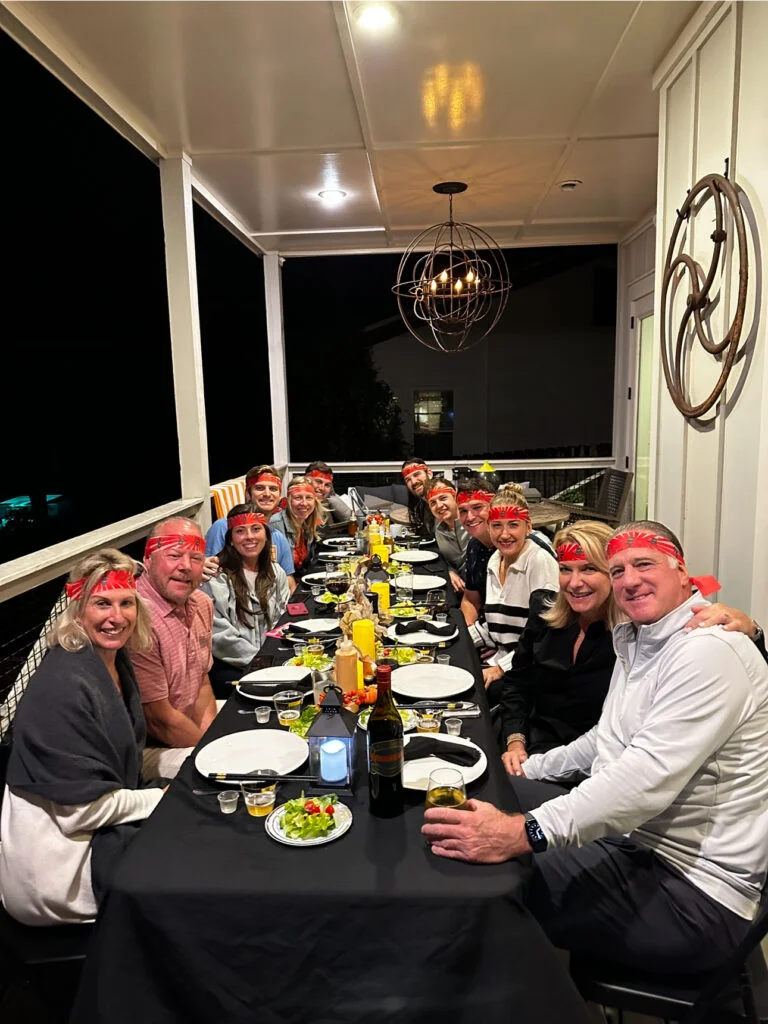 A large group of people enjoying a hibachi dinner at a long, elegantly set table on a cozy outdoor porch at night. Everyone is smiling, wearing matching red bandanas, and surrounded by plates of food, drinks, and decorative lighting.