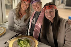 Three women enjoying a lively hibachi dinner party at Alys Beach, seated at a beautifully set table with gold-rimmed plates and fresh salad, celebrating with festive headbands